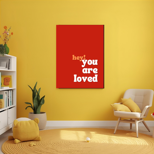 Red wall art with 'hey you are loved' text on a yellow wall in a room with a chair and books.
